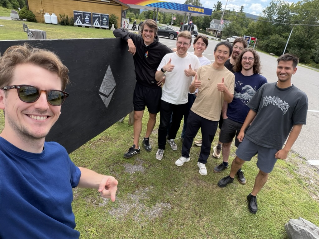 The Ethlabs team giving thumbs up around the black wooden board with a carved Ethereum logo, in front of a gas station in Hunter, NY